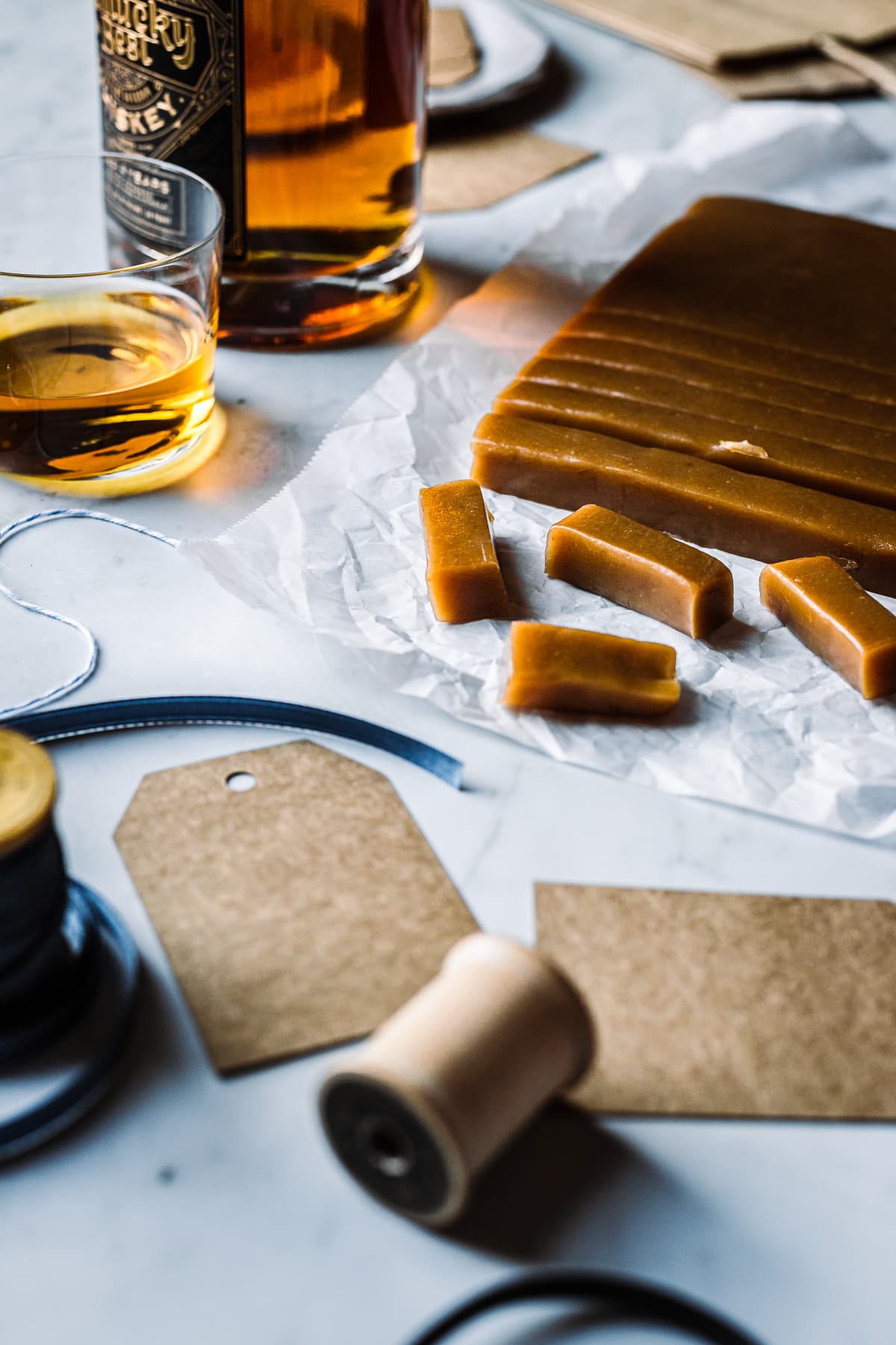 Candy on white parchment paper being cut into individual pieces, surrounded by gift wrapping supplies and a bottle of whiskey.