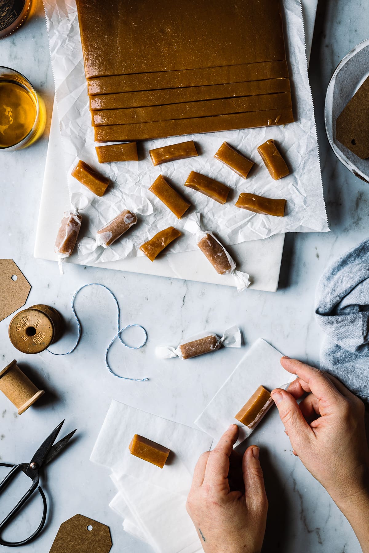 A batch of candy being cut into individual pieces and wrapped. The work surface is grey marble. There are paper candy wrappers, a vintage spool of twine, and scissors nearby. White hands reach into the frame to wrap a piece of candy in paper.
