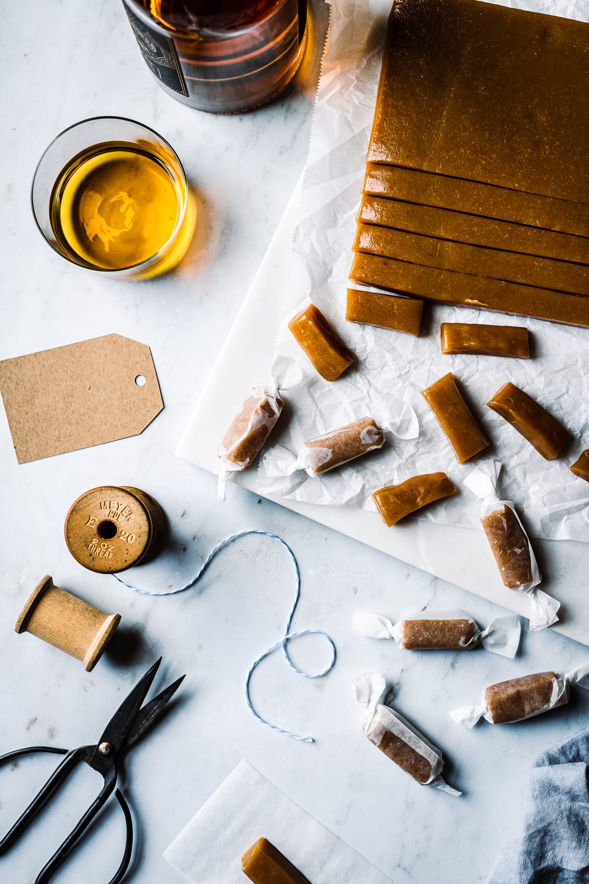 Pieces of candy being cut into individual pieces and wrapped. The work surface is grey marble. There are brown paper gift tags, a vintage spool of twine, scissors and a glass of whiskey nearby. A bottle of whiskey peeks into the frame at top left.