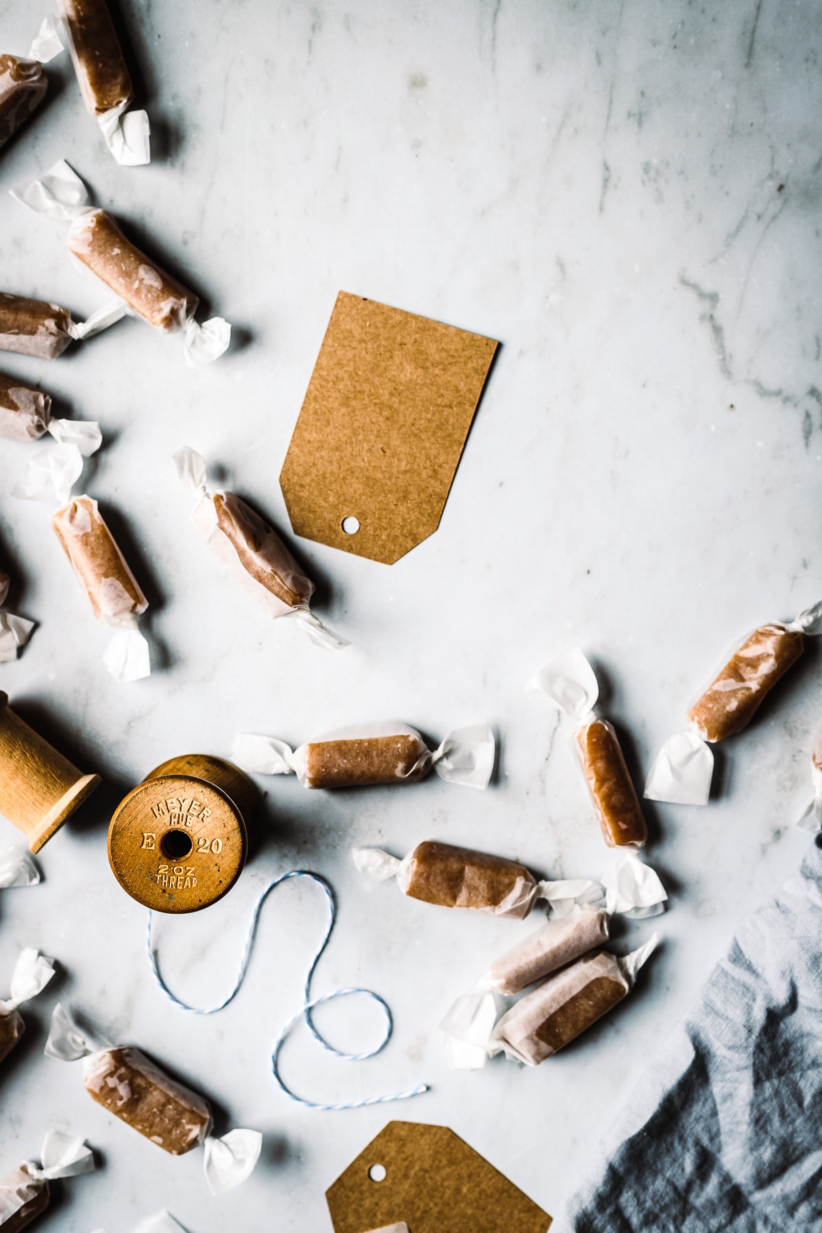 Individually wrapped whiskey salted caramels on a grey marble surface. There is a vintage spool of twine, brown paper gift tags, and a blue linen napkin nearby.