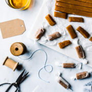 Pieces of candy being cut into individual pieces and wrapped. The work surface is grey marble. There are brown paper gift tags, a vintage spool of twine, and scissors nearby. A glass of whiskey peeks into the frame at top left.