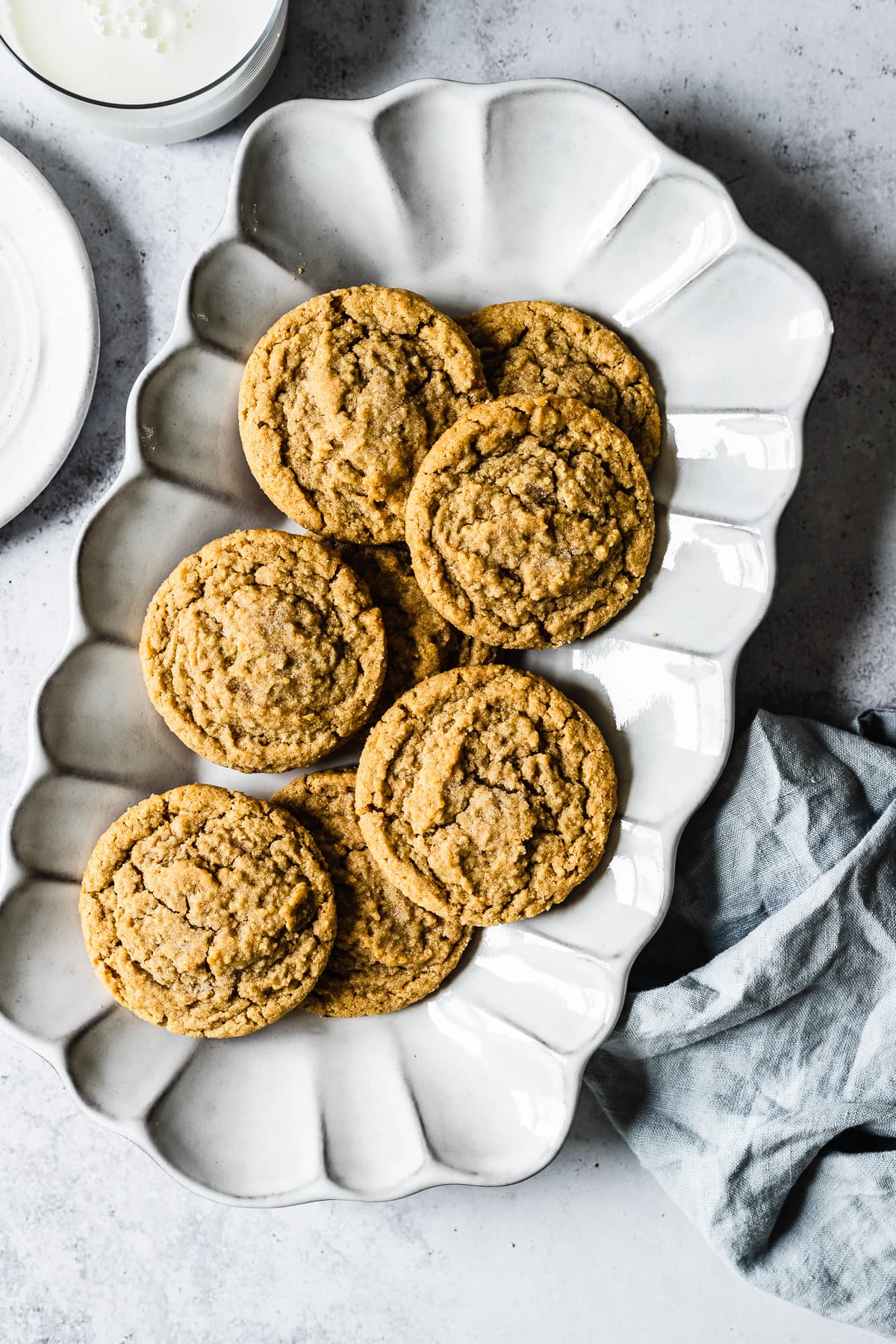 Peanut butter cookies on a fluted white ceramic serving plate. The plate rests on a white stone surface with a light blue linen napkin, a white plate, and a glass of milk nearby.