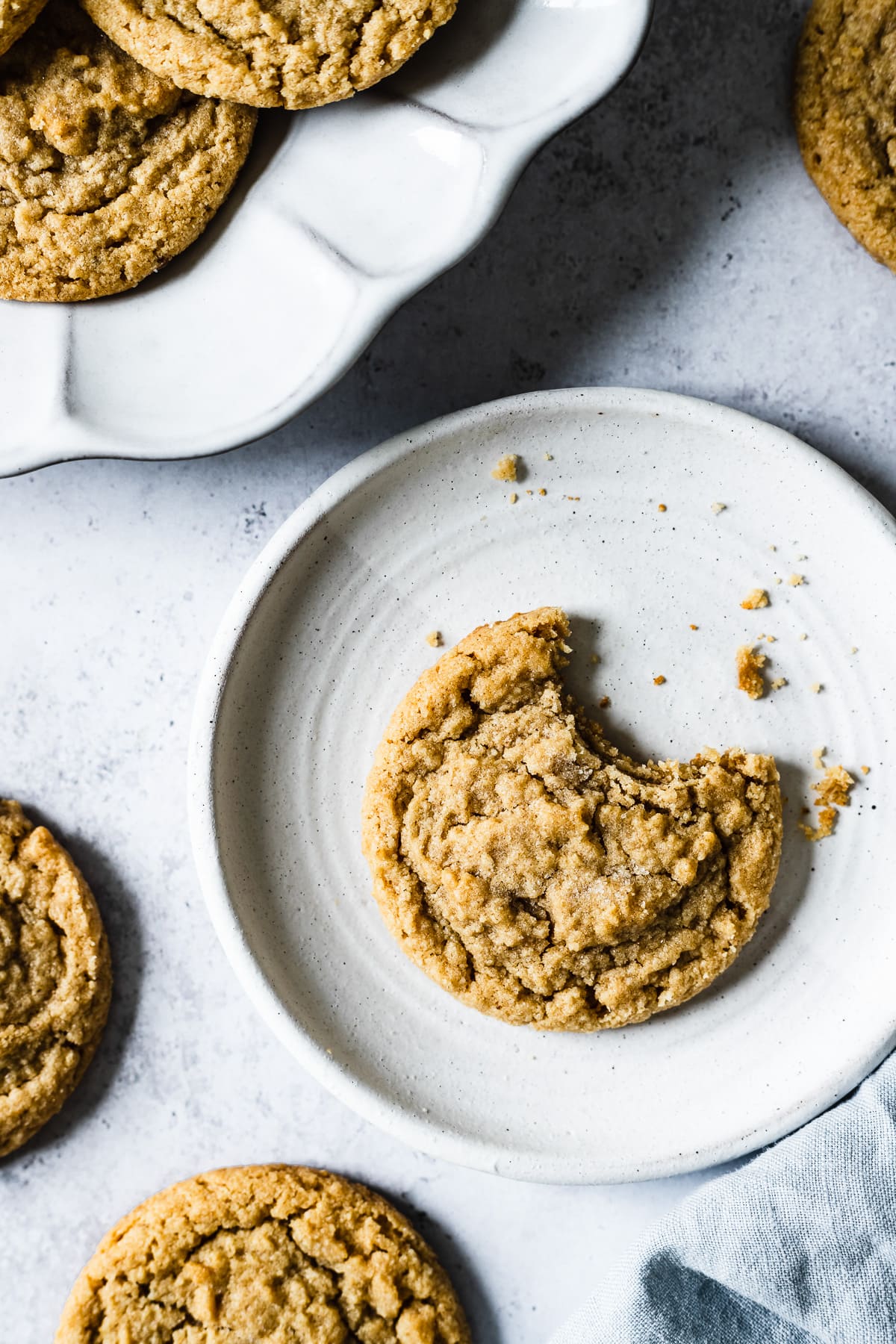 A cookie with a bite taken out of it on a round white cermic plate. More cookies and a light blue linen napkin are nearby, as well as a fluted ceramic serving plate with more cookies.