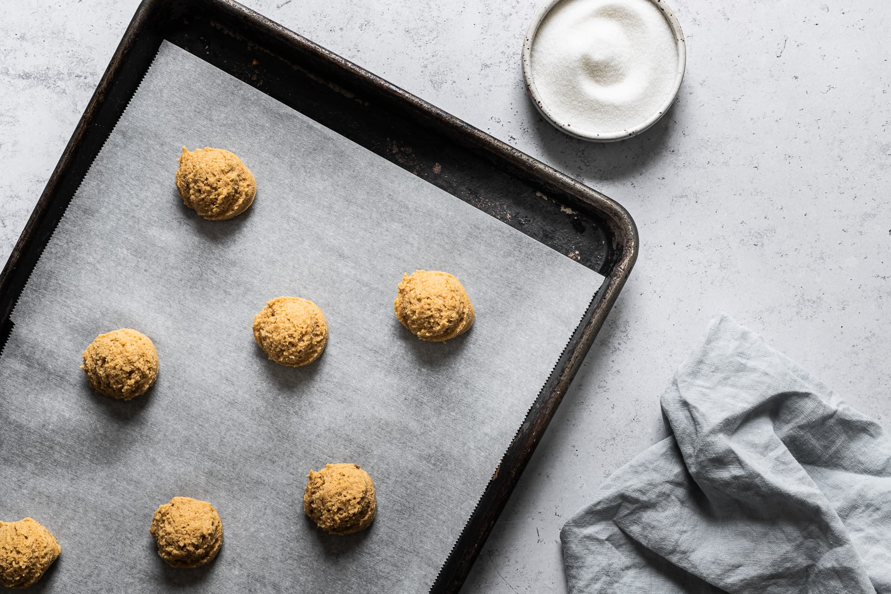 Scoops of almond flour peanut butter cookie dough on a parchment lined baking sheet with a small bowl of granulated sugar nearby.