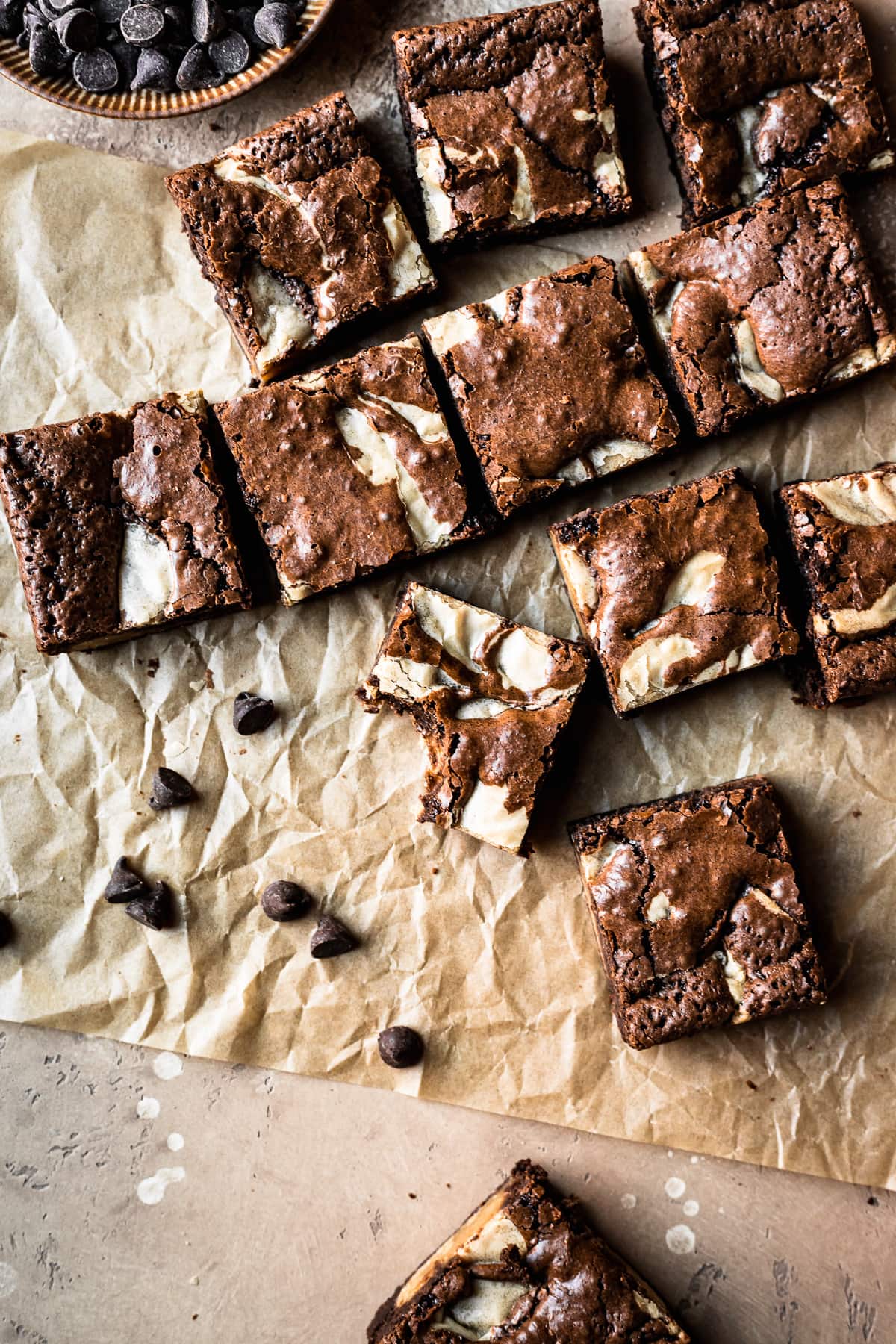 Sliced squares of brownies casually arranged on brown parchment underneath. A bowl of chocolate chips peeks into the frame at top left. The background surface is textured tan stone.