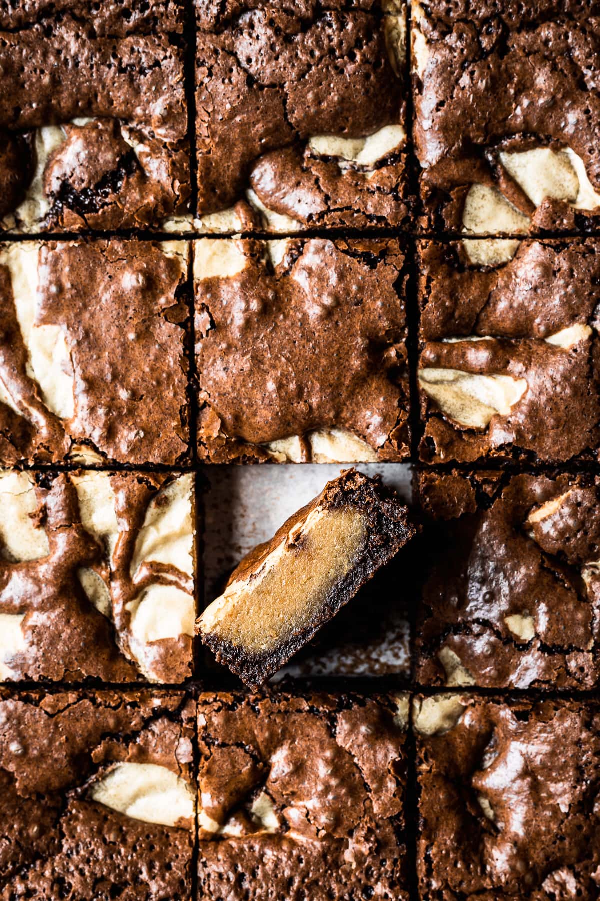 A closeup of a pan of brownie blondies sliced into squares with light and dark swirls thoughout in a marble pattern. The crust has a shiny, crackly top. One square has been turned on edge to show the interior.