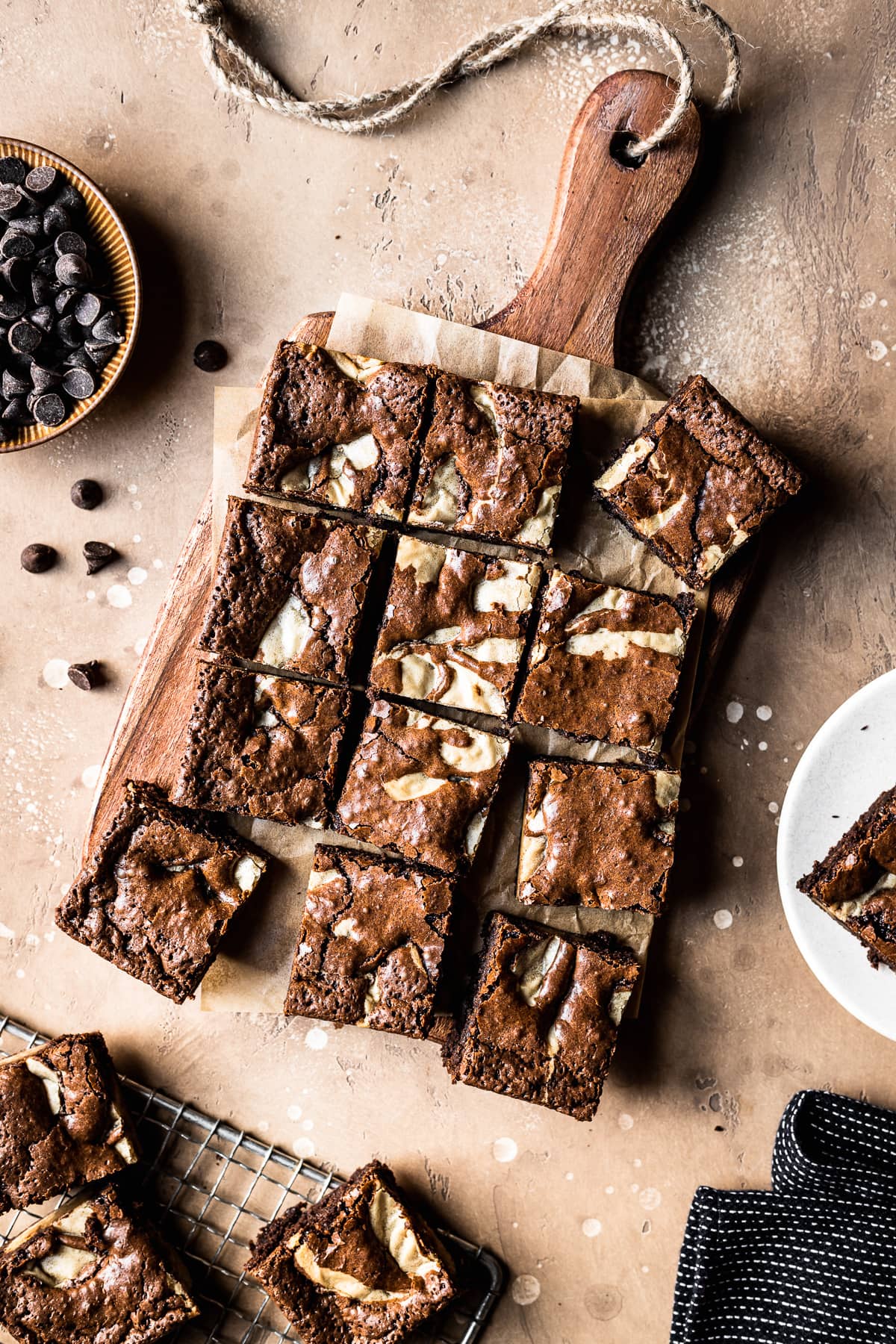 Brownie blondie squares casually arranged on brown parchment paper with a wooden cutting board underneath. A bowl of chocolate chips, a small plate with a brownie on top, and a dark blue napkin peek into the frame. The background surface is textured tan stone.