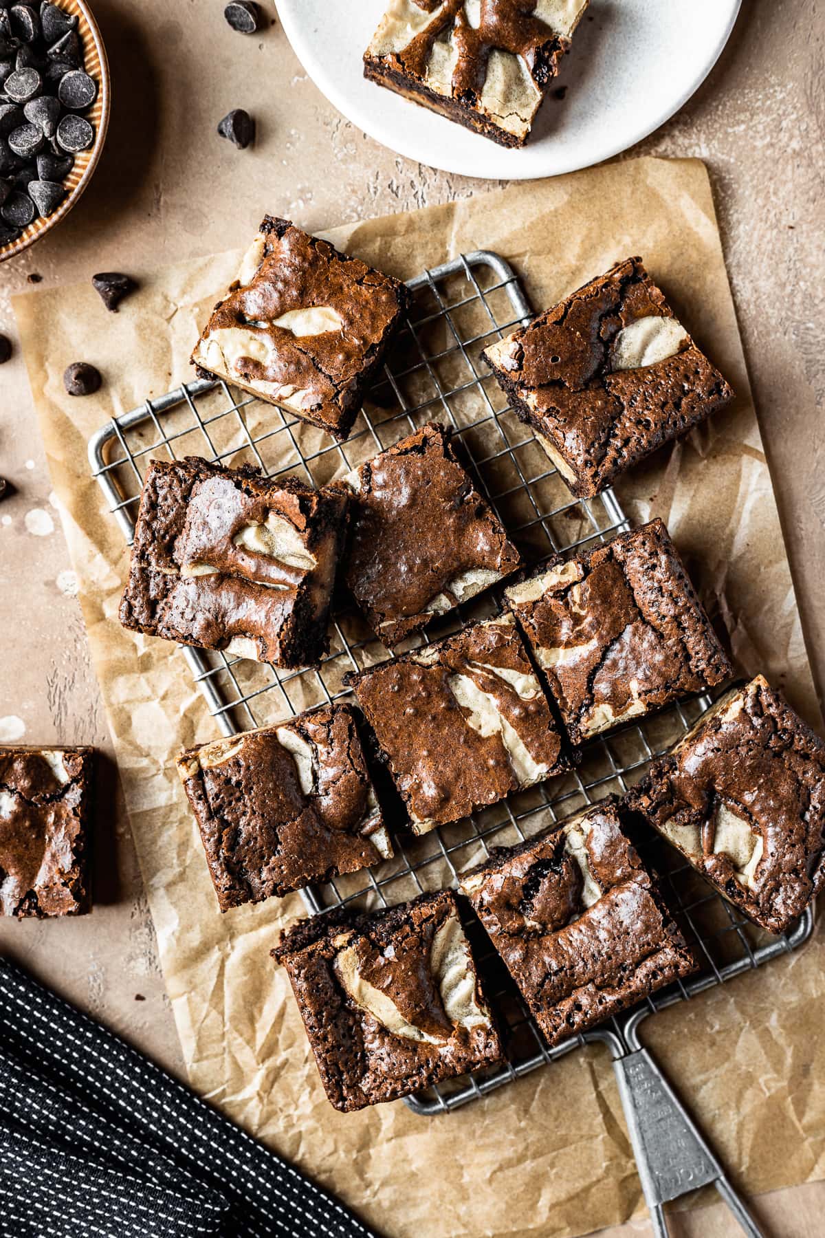 Brownie blondie squares casually arranged on a cooling rack with brown parchment underneath. A bowl of chocolate chips, a small plate with a brownie on top, and a dark blue napkin peek into the frame. The background surface is textured tan stone.