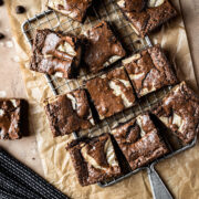 Brownie blondie squares casually arranged on a cooling rack with brown parchment underneath. A bowl of chocolate chips, a small plate with a brownie on top, and a dark blue napkin peek into the frame. The background surface is textured tan stone.