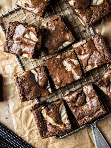 Brownie blondie squares casually arranged on a cooling rack with brown parchment underneath. A bowl of chocolate chips, a small plate with a brownie on top, and a dark blue napkin peek into the frame. The background surface is textured tan stone.