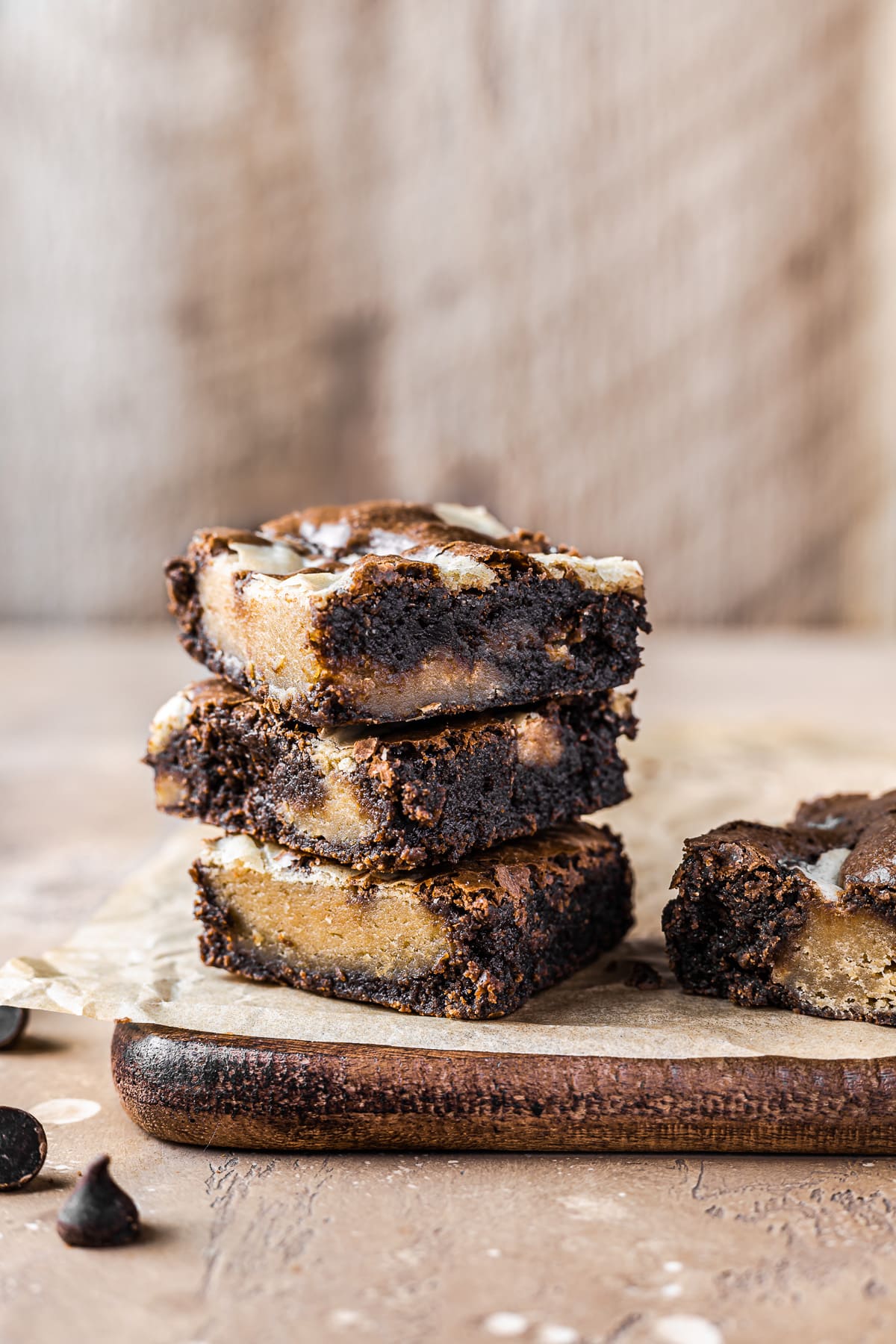 Three brownie blondie bars stacked on top of each other on a wooden cutting board. Another bar rests to the right. A warm tan textured surface is underneath. The background is also a warm tan textured pattern.