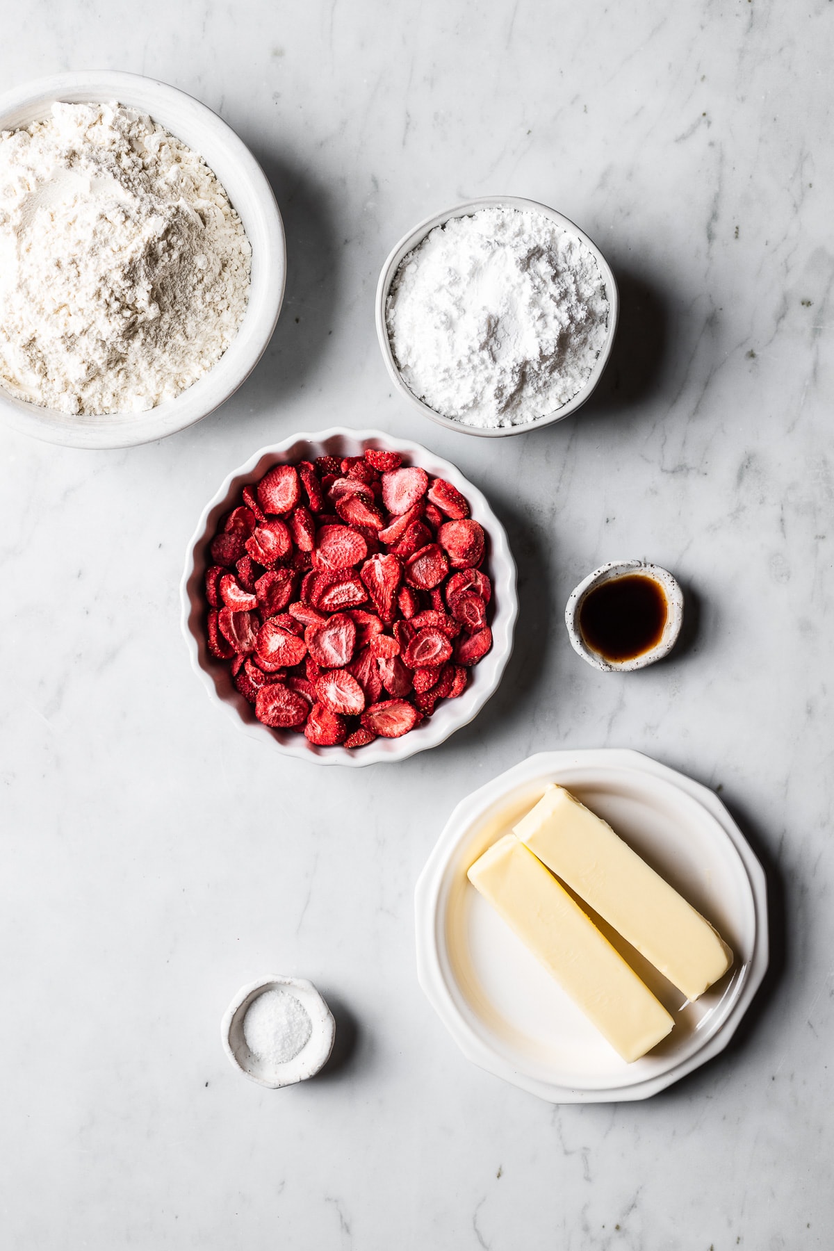 A photo of ingredients for strawberry shortbread cookies in white ceramic conainers on a light grey marble surface.