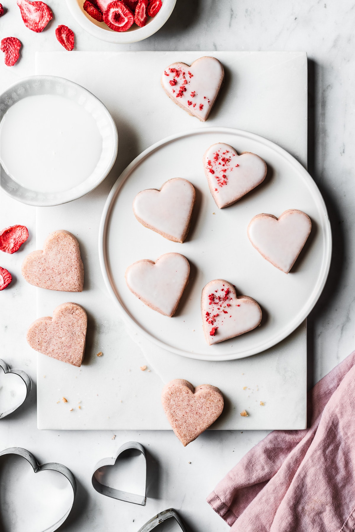 Heart shaped cookies on a white plate and a white marble tray on a light grey marble surface. Some of the cookies have been dipped in white glaze. Heart shaped cookie cutters, a bowl of freeze dried strawberries, and a pink napkin rest nearby.