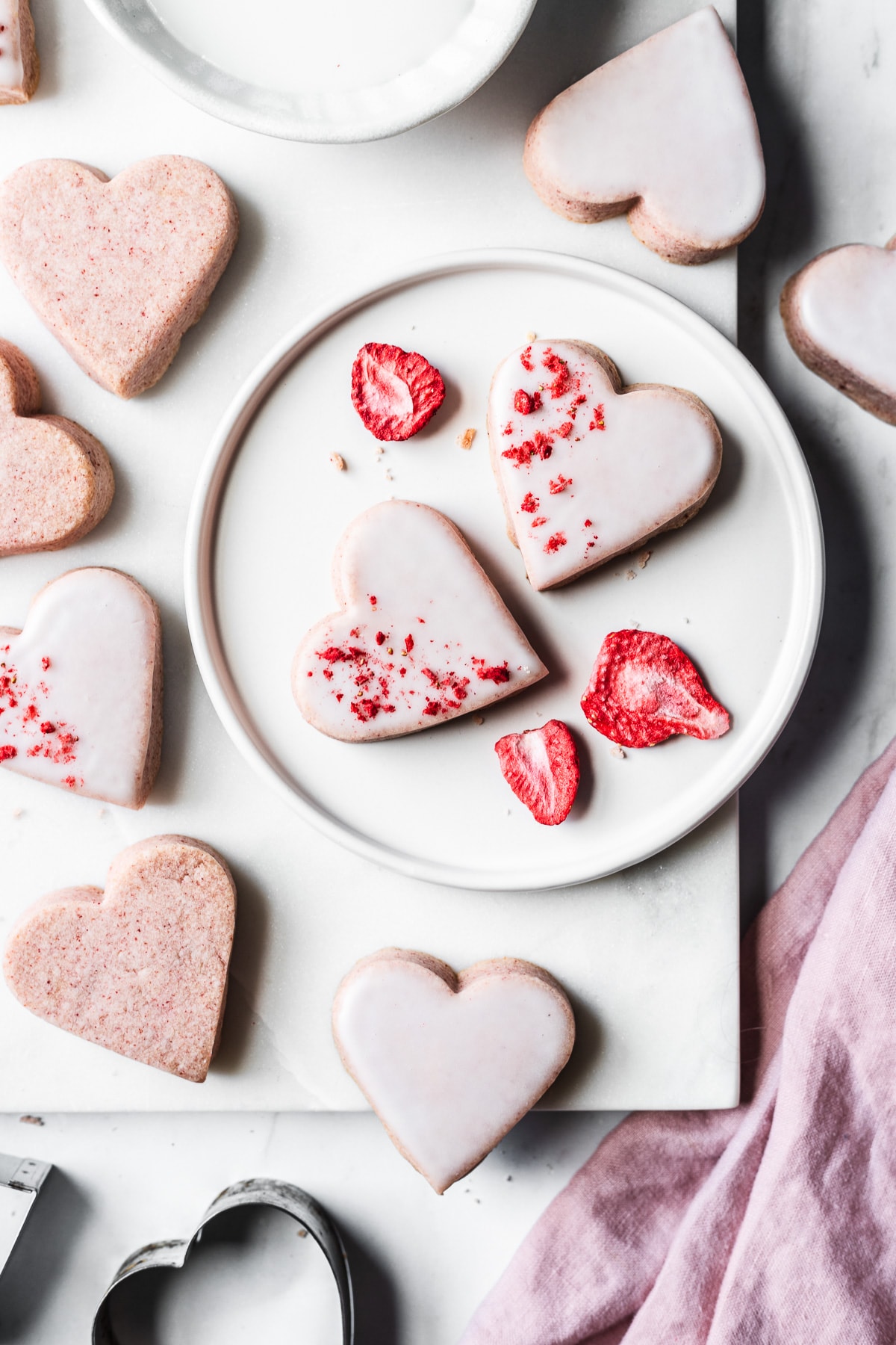 Glazed heart shaped cookies on a white plate and a white marble tray on a light grey marble surface. Some of the cookies have been dipped in white glaze. Heart shaped cookie cutters, a white ceramic bowl of glaze, and a pink napkin rest nearby.