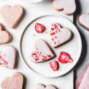 Glazed heart shaped cookies on a white plate and a white marble tray on a light grey marble surface. Some of the cookies have been dipped in white glaze and sprinkled with red freeze dried strawberries. A pink napkin peeks into the frame at bottom right.