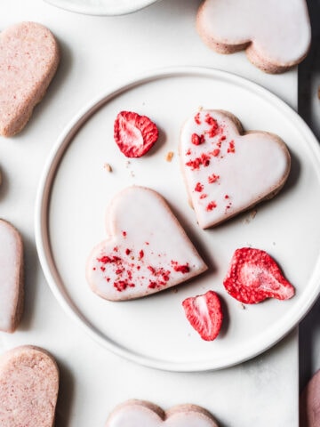 Glazed heart shaped cookies on a white plate and a white marble tray on a light grey marble surface. Some of the cookies have been dipped in white glaze and sprinkled with red freeze dried strawberries. A pink napkin peeks into the frame at bottom right.