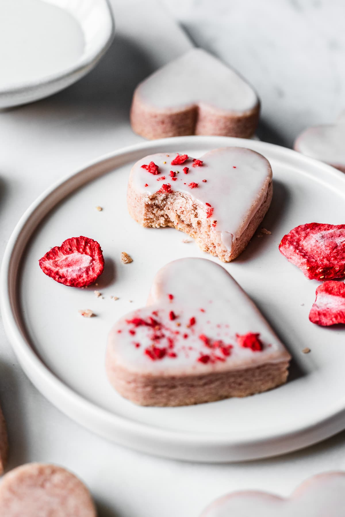 Glazed heart shaped cookies on a white plate and a white marble tray on a light grey marble surface. Some of the cookies have been sprinkled with red freeze dried strawberries. One of the cookies has a bite taken out of it, revealing the crumb.