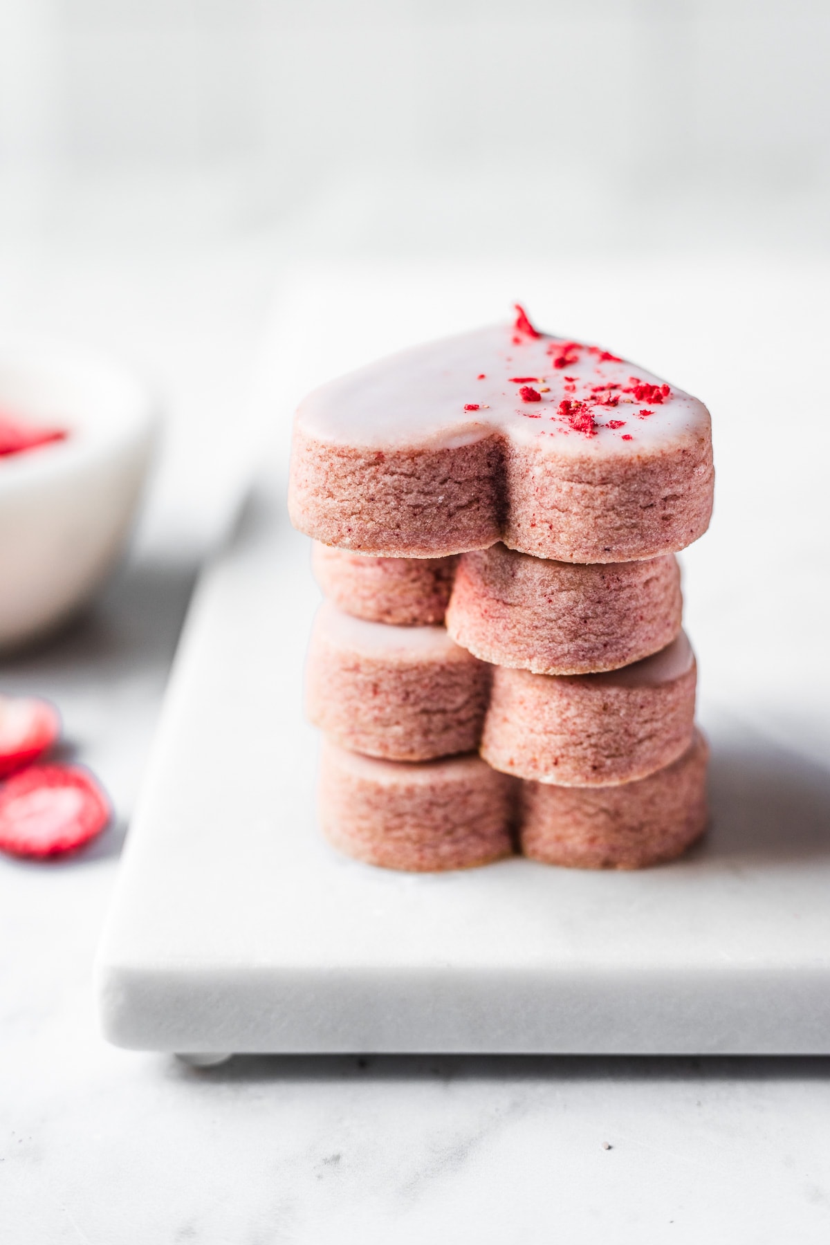 A side view of a stack of four glazed pink heart shaped cookies on a marble tray. The image is light and bright. A white bowl of freeze dried strawberries peeks into the frame at left.