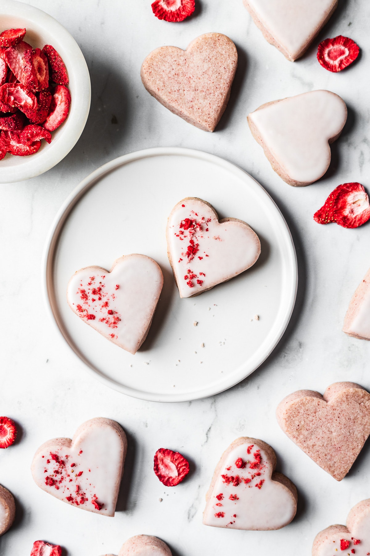Two glazed heart shaped strawberry shortbread cookies on a white plate on a light grey marble surface, surrounded by more glazed and unglazed cookies. A bowl of freeze dried strawberries is at top left. Some of the cookies have been dipped in white glaze and sprinkled with red freeze dried strawberries.
