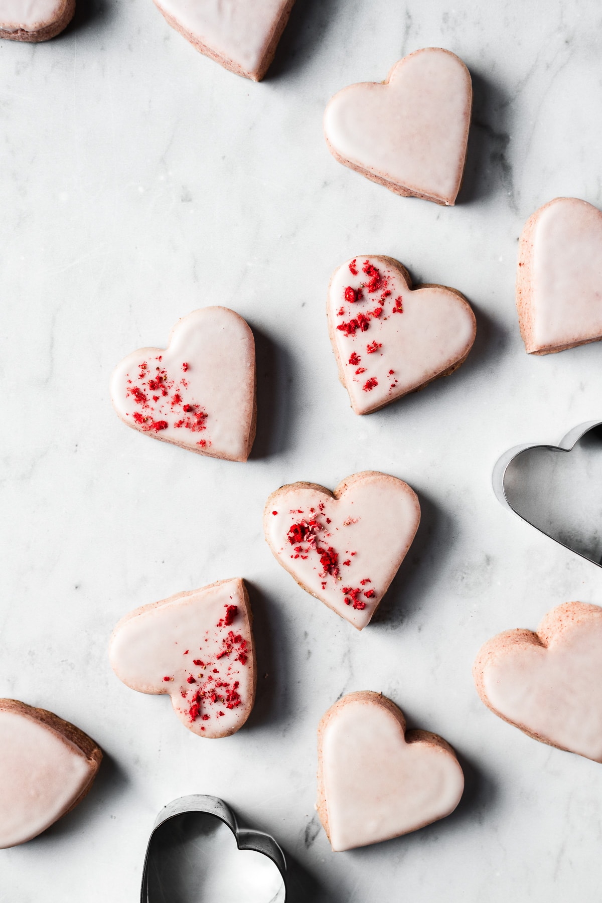 Glazed pink heart shaped strawberry shortbread cookies on a light grey marble surface. Some of the cookies are sprinkled with red freeze dried strawberries.