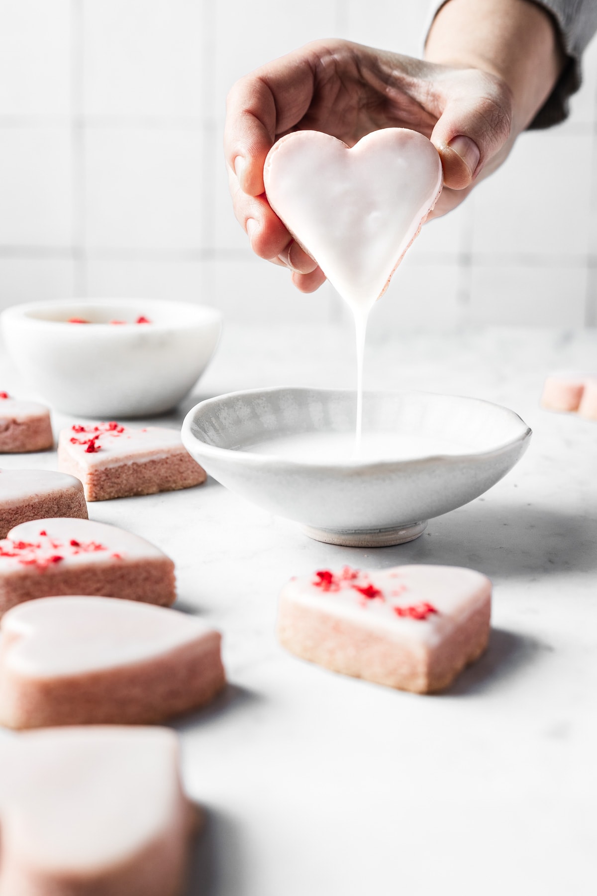 A hand dips a pink shortbread cookie into a white ceramic bowl of glaze. The extra glaze drips back into the bowl. Glazed cookies rest on the marble surface around the bowl. Some have been sprinkled with red freeze dried strawberries.