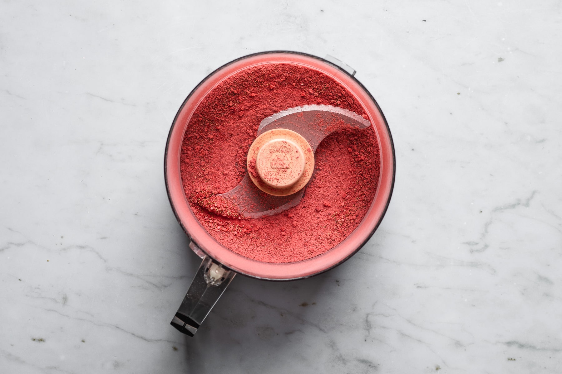 A process photo of ground up freeze dried strawberries in a food processor on a light grey marble surface.