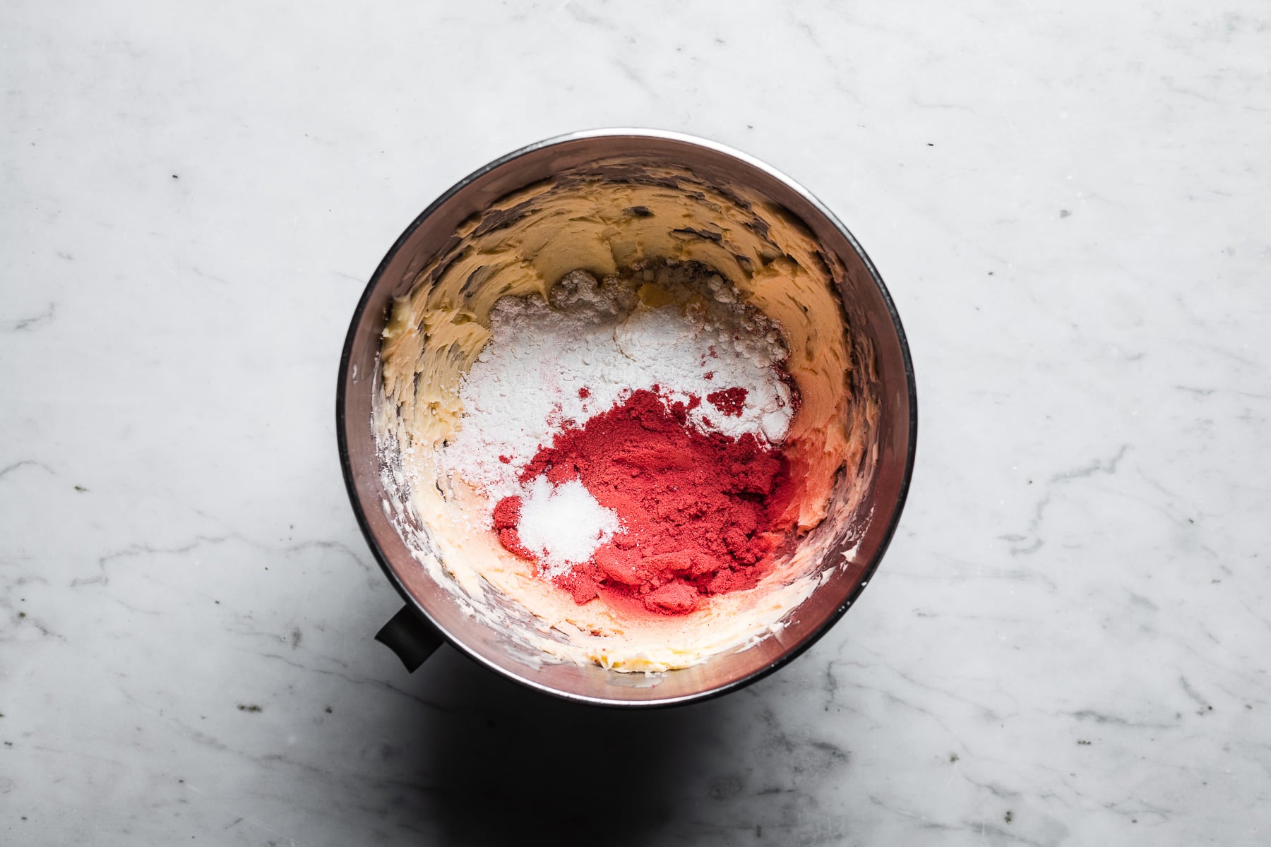 A process photo showing butter and dry ingredients in a metal mixing bowl, resting on a light grey marble surface.