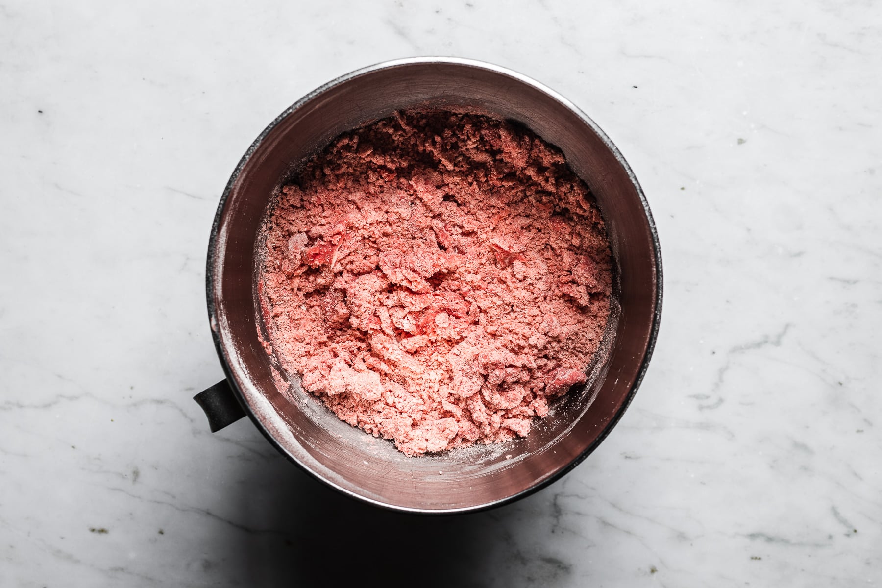 A process photo showing cookie batter in a metal mixing bowl, resting on a light grey marble surface.