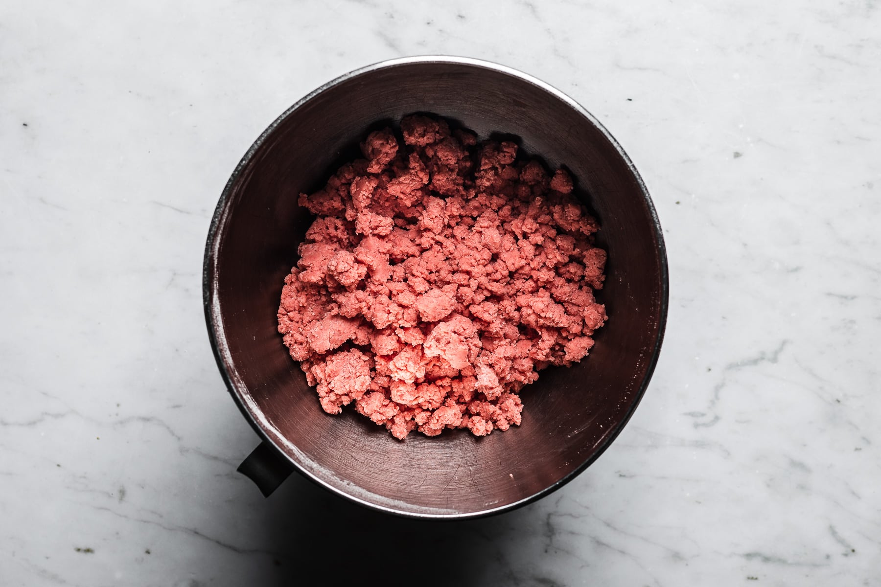 A process photo showing pink shortbread dough in a metal mixing bowl, resting on a light grey marble surface.