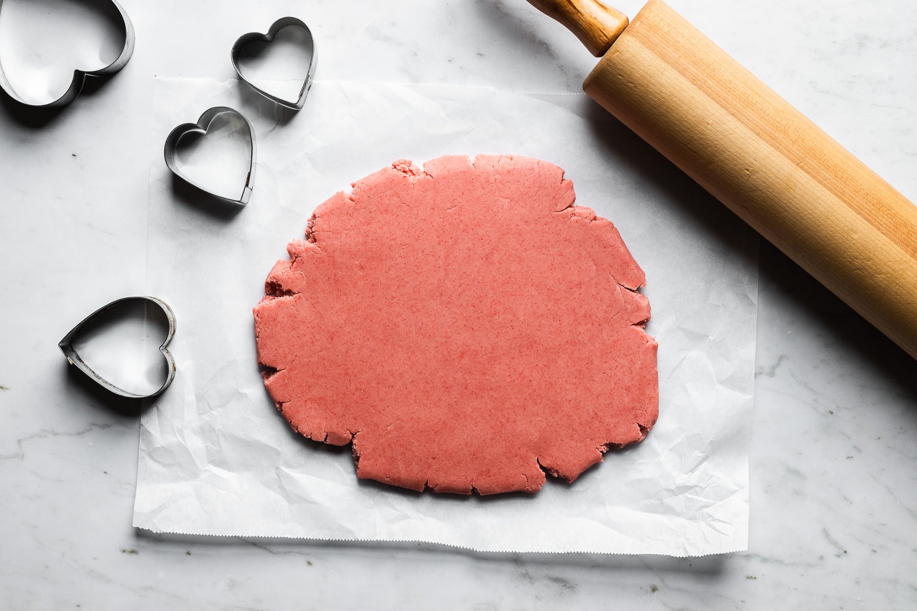 PInk shortbread dough rolled out on a rectangle of white parchment paper on a grey marble surface. Heart shaped cookie cutters and a rolling pin rest nearby.