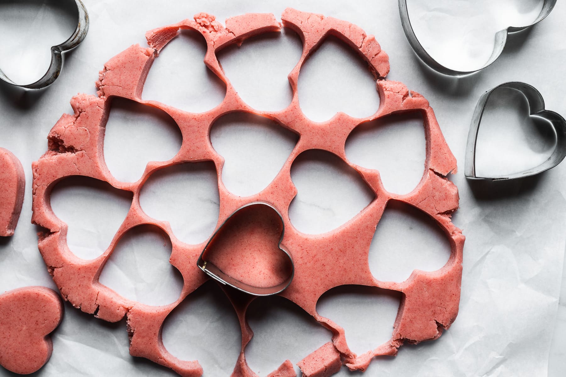 Pink cookie dough rolled out on white parchment paper with heart shaped holes where the dough has been removed. Heart shaped metal cookie cutters rest nearby.