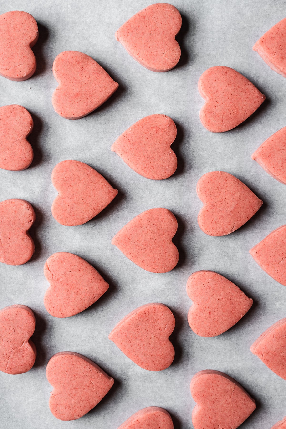 Rows of unbaked pink heart shaped cookies on a white parchment surface.