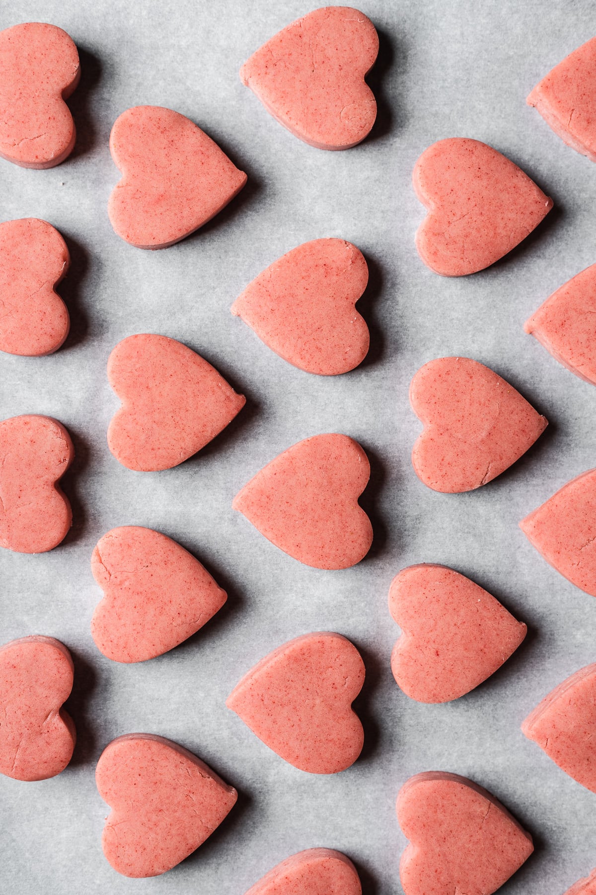 Rows of unbaked pink heart shaped cookies on white parchment paper.