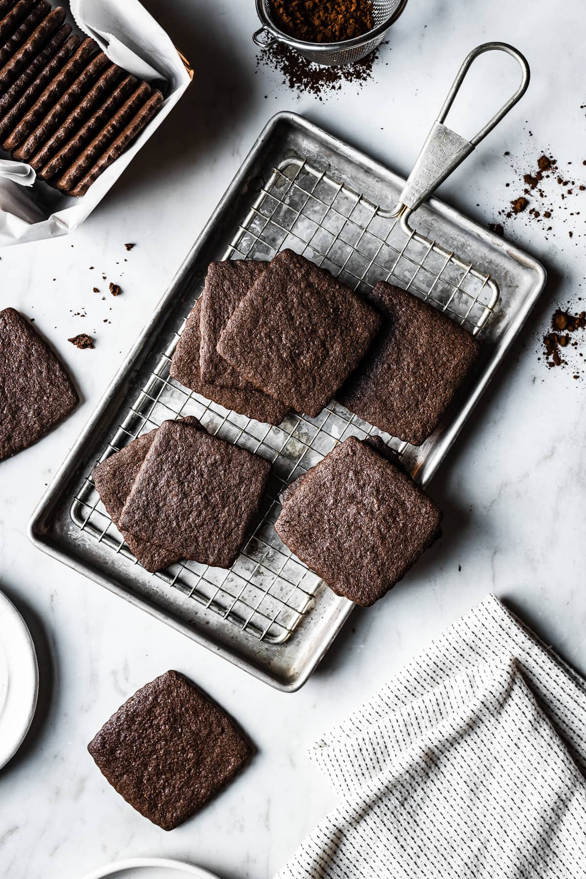 Chocolate wafer cookies on a vintage metal baking sheet and cooling rack on a marble surface. A container with a stack of cookies in it peeks into the image top left, along with a metal sieve of cocoa powder. There are cookie crumbs and cocoa powder scattered on the marble surface.
