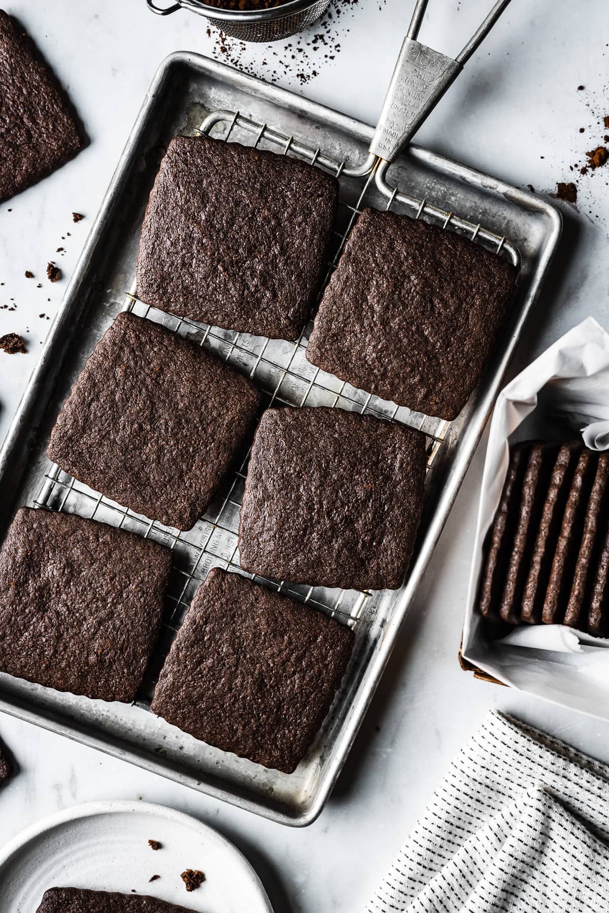 Chocolate wafer cookies on a vintage metal baking sheet and cooling rack on a marble surface. A container with a stack of cookies in it peeks into the image at right. There are cookie crumbs and cocoa powder on the marble surface.