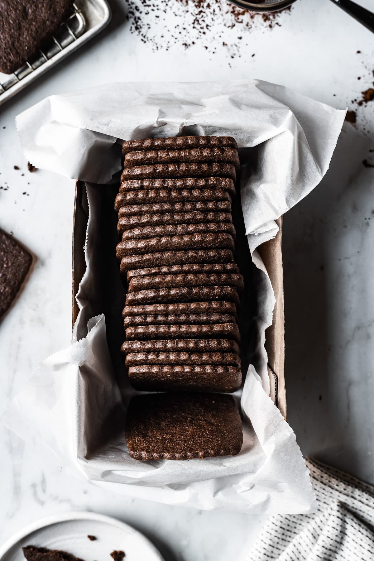 Square homemade chocolate wafer cookies in a white parchment paper lined box on a marble surface. A baking rack, cookies, a white ceramic plate and a white napkin peek into the image from the edges.