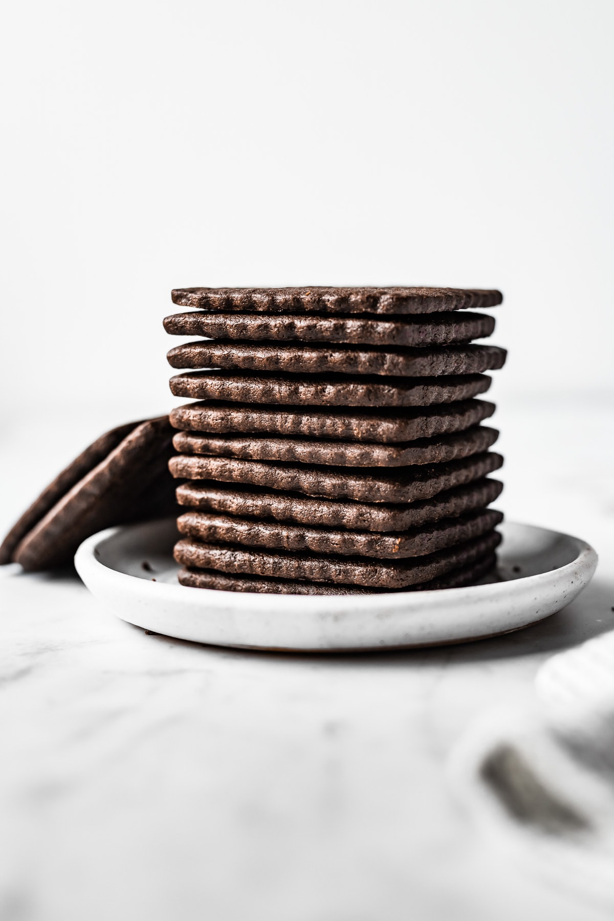A stack of square chocolate cookies with scalloped edge on a small white ceramic plate on a light grey marble surface. Two additional cookies on the left lean against the stack.