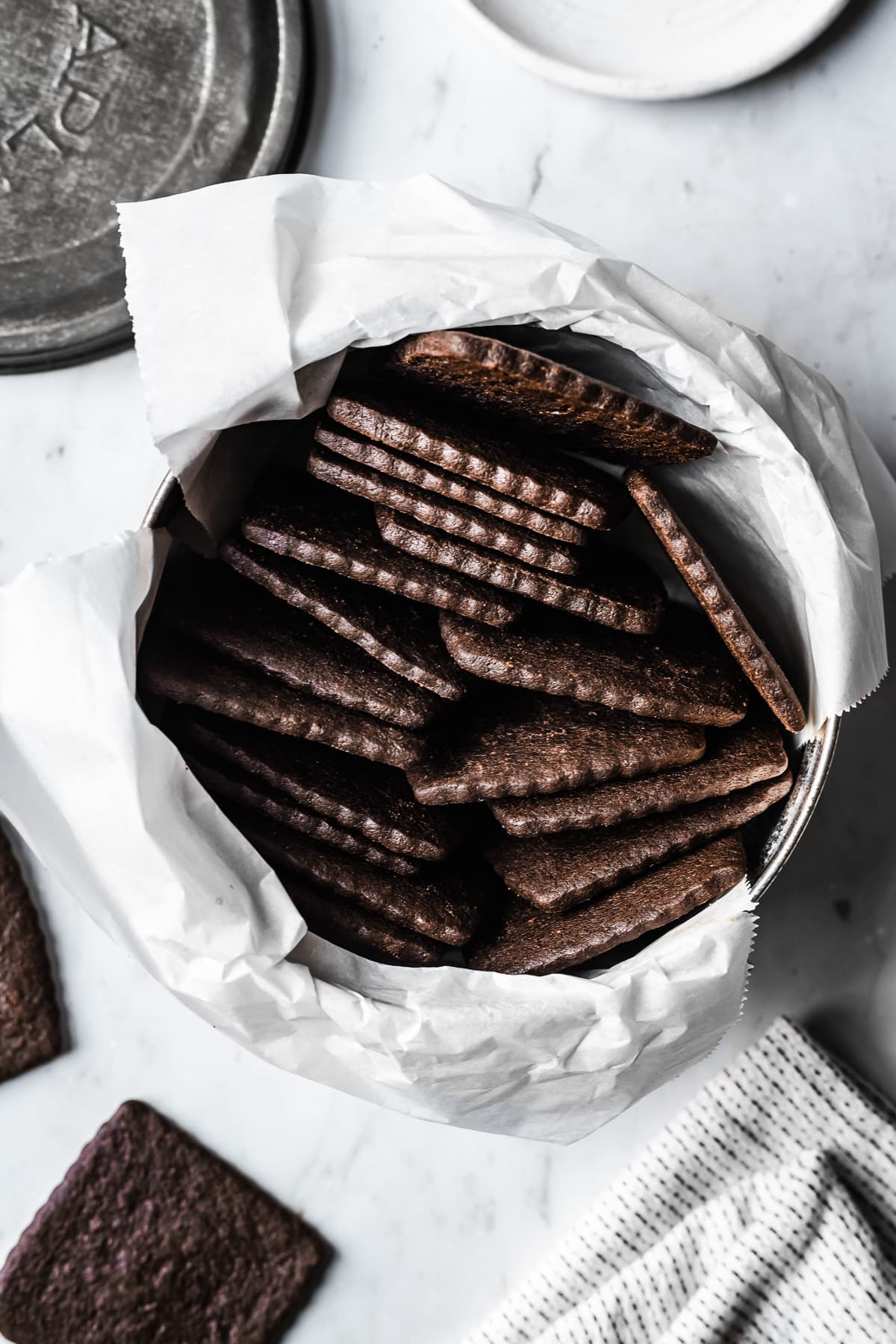 Chocolate wafer cookies in a parchment lined vintage metal tin on a light grey marble surface. Several cookies, a white striped napkin, and a ceramic plate peek into the image.