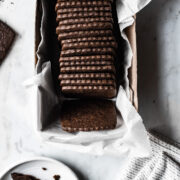 Square homemade chocolate wafer cookies in a white parchment paper lined box on a marble surface. A cookie, a white ceramic plate and a white napkin peek into the image from the edges.