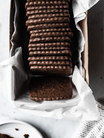 Square homemade chocolate wafer cookies in a white parchment paper lined box on a marble surface. A cookie, a white ceramic plate and a white napkin peek into the image from the edges.