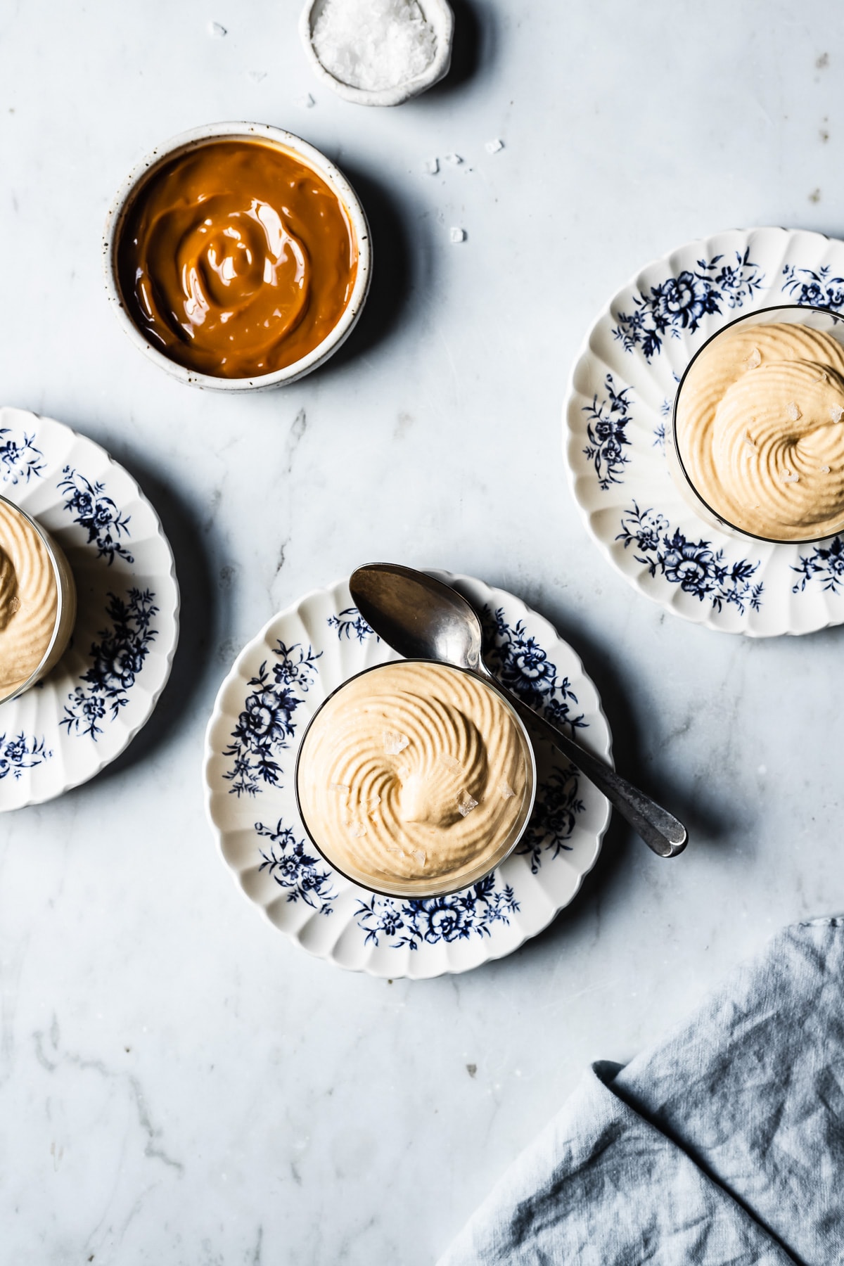 Three thin glass cups hold circular swirls of piped dulce de leche mousse. The cups sit on small scalloped blue and white floral plates on a grey marble background. Nearby are small bowls of dulce de leche and flaky sea salt. A light blue linen napkin rests at bottom right.