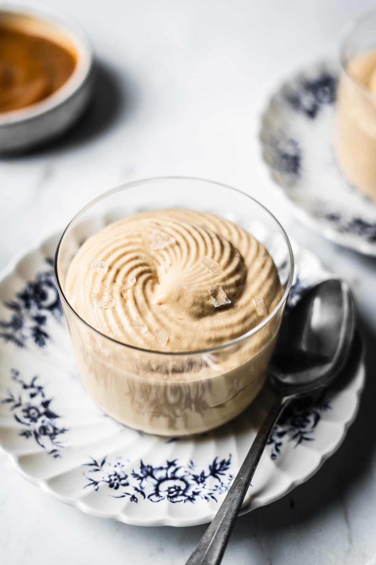 A thin glass cup holds circular swirls of piped dulce de leche mousse with flaky sea salt on top. The cup sits on a small scalloped blue and white floral plate with a spoon resting on it. The surface is made of grey marble background. At left, out of focus, is a small bowl of dulce de leche.