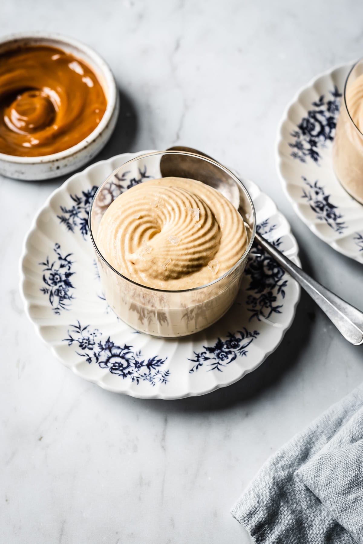 A thin glass cup holds circular swirls of piped dulce de leche mousse with flaky sea salt on top. The cup sits on a small scalloped blue and white floral plate with a spoon resting on it. The surface is made of grey marble background. At left, out of focus, is a small bowl of dulce de leche. Another glass of mousse peeks into the frame at right, along with a light blue linen napkin.