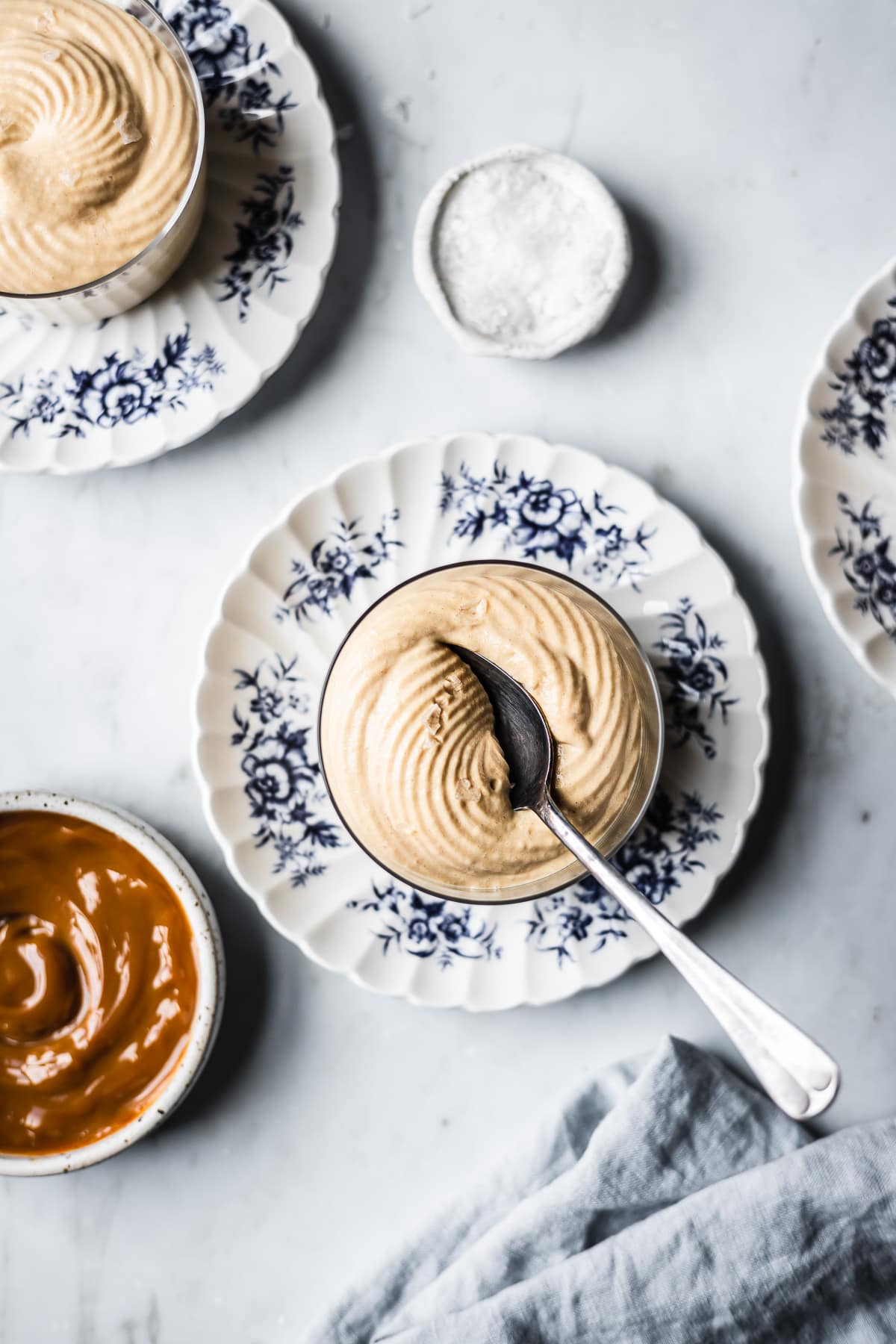 Two thin glass cups hold circular swirls of piped dulce de leche mousse. The cups sit on small scalloped blue and white ceramic floral plates on a grey marble background. Nearby are small bowls of dulce de leche and flaky sea salt. A light blue linen napkin rests at bottom right.