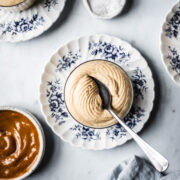 A thin glass cup holds circular swirls of piped dulce de leche mousse with a spoon resting in it. The cup sits on a small scalloped blue and white ceramic floral plate on a light grey marble background. Nearby are small bowls of dulce de leche and flaky sea salt. A light blue linen napkin peeks into the frame at bottom right.