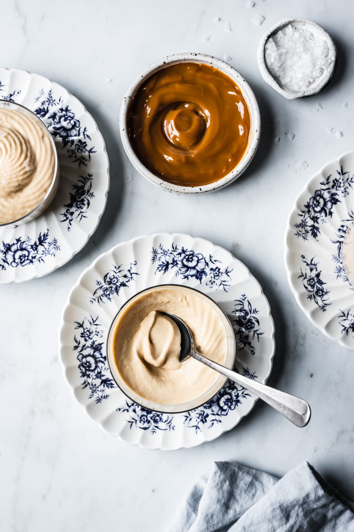 Three thin glass cups hold circular swirls of piped dulce de leche mousse. The cups sit on small scalloped blue and white floral plates on a grey marble background. Nearby are small bowls of dulce de leche and flaky sea salt. A light blue linen napkin rests at bottom right.