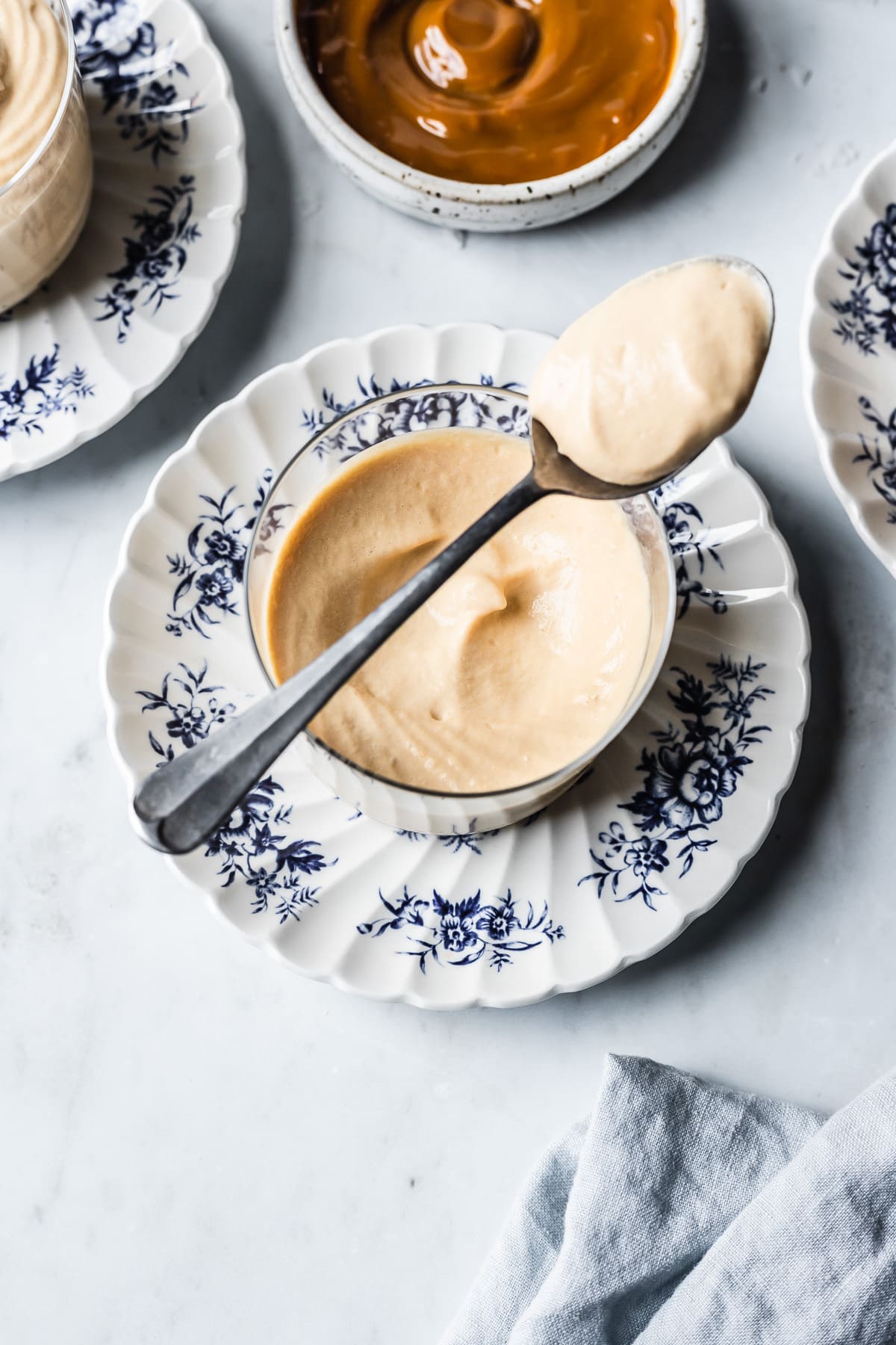 A thin glass cup holds circular swirls of piped dulce de leche mousse. A spoon with a scoop full of mousse rests on top of the glass. The cup sits on a small scalloped blue and white ceramic floral plate on a light grey marble background. At top is a small white ceramic bowl of dulce de leche. A light blue linen napkin peeks into the frame at bottom right.