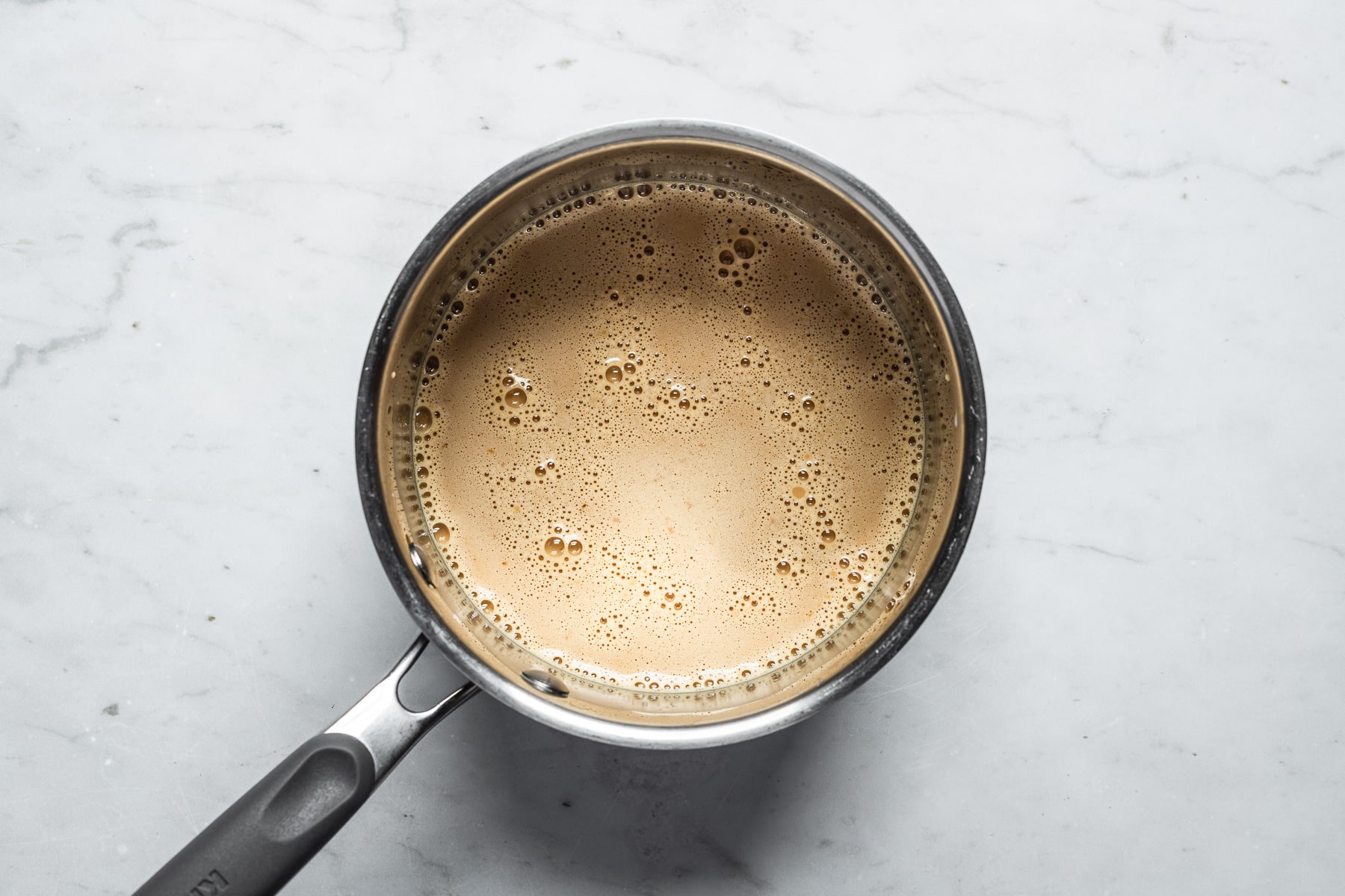 A process photo showing a metal saucepan of milk and cornstarch added to the dulce de leche to make pastry cream. The saucepan sits on a grey marble surface.