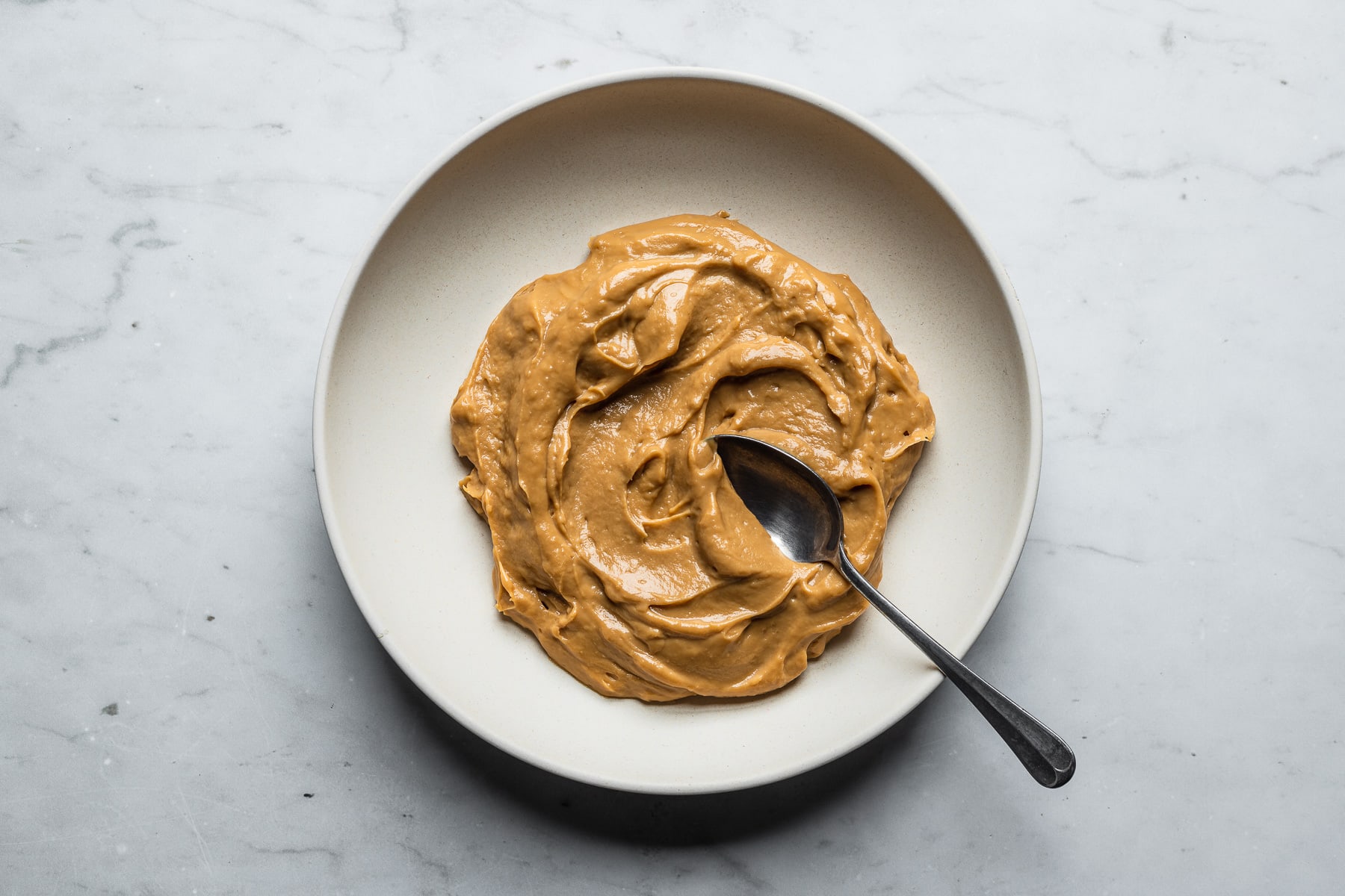 A process photo showing cooked dulce de leche pastry cream in a tan bowl with a spoon in it. The bowl sits on a marble surface.