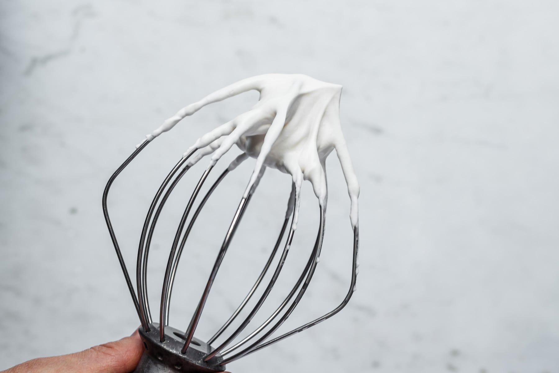 A process photo showing the wire whisk from a stand mixer holding whipped cream beaten to soft peaks.